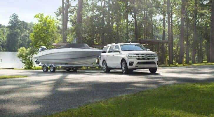 A white 2024 Ford Expedition is shown towing a boat near a lake.