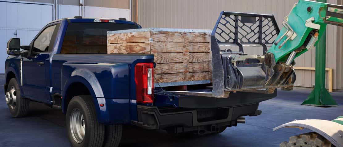 A blue Ford Super Duty having cargo loaded into its truck bed by a green crane.