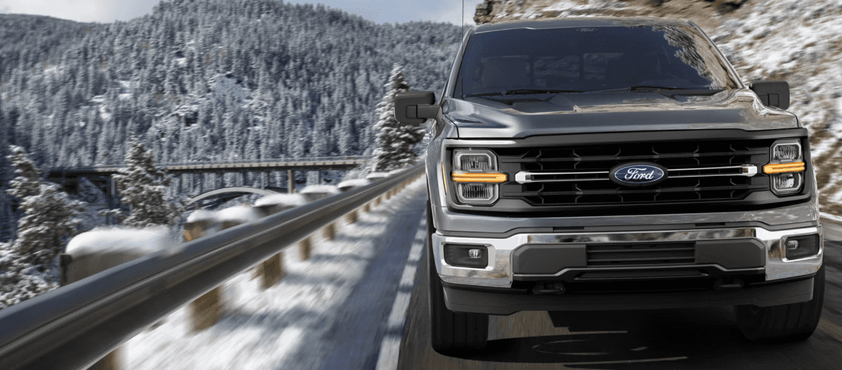 A gray Ford F-150 driving on a road by a cliff in a snowy area.ALT