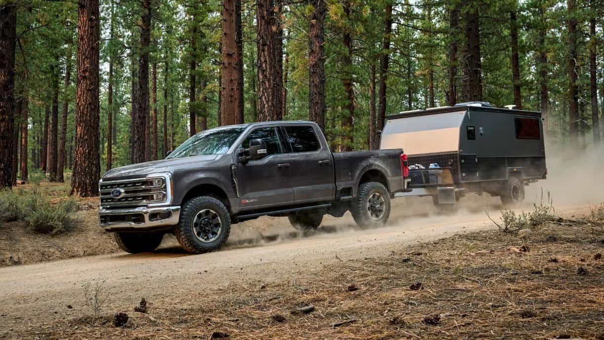 A gray Ford F-250 towing a small trailer over a dirt road in a forest.