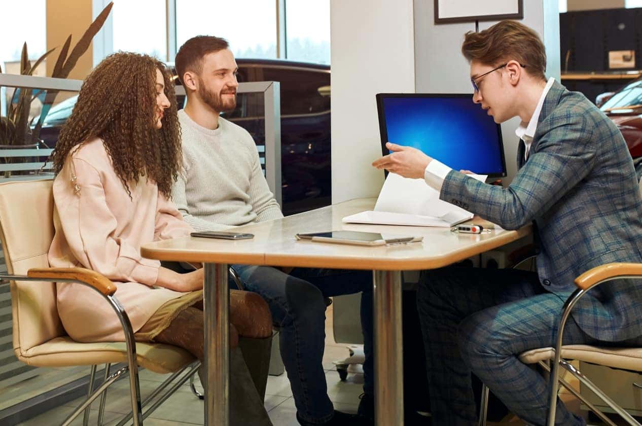 A couple sits across from a man in a suit at a dealership desk. They're engaged in discussion, papers on the table, and a computer screen nearby.