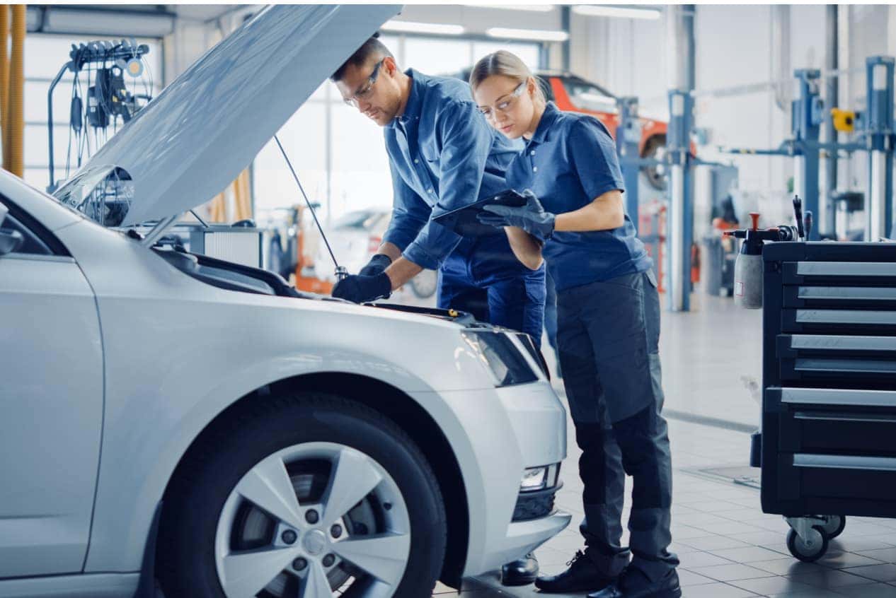 Two mechanics in a garage examine a car engine, wearing protective eyewear and gloves.