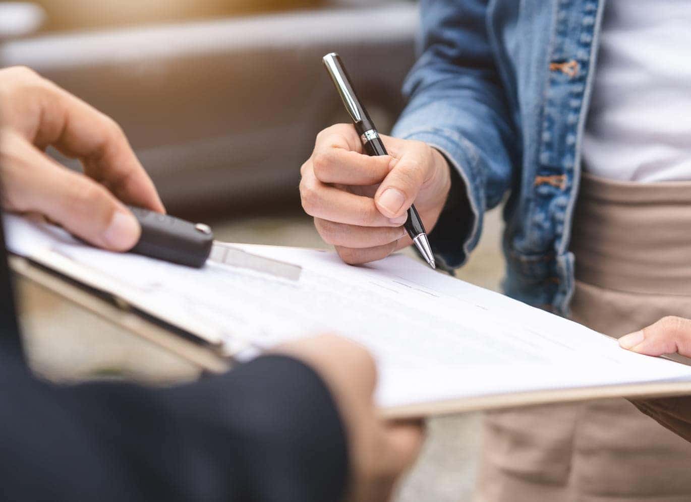 A close-up of a customer signing paperwork on a clipboard with a pen while another person holds the documents.