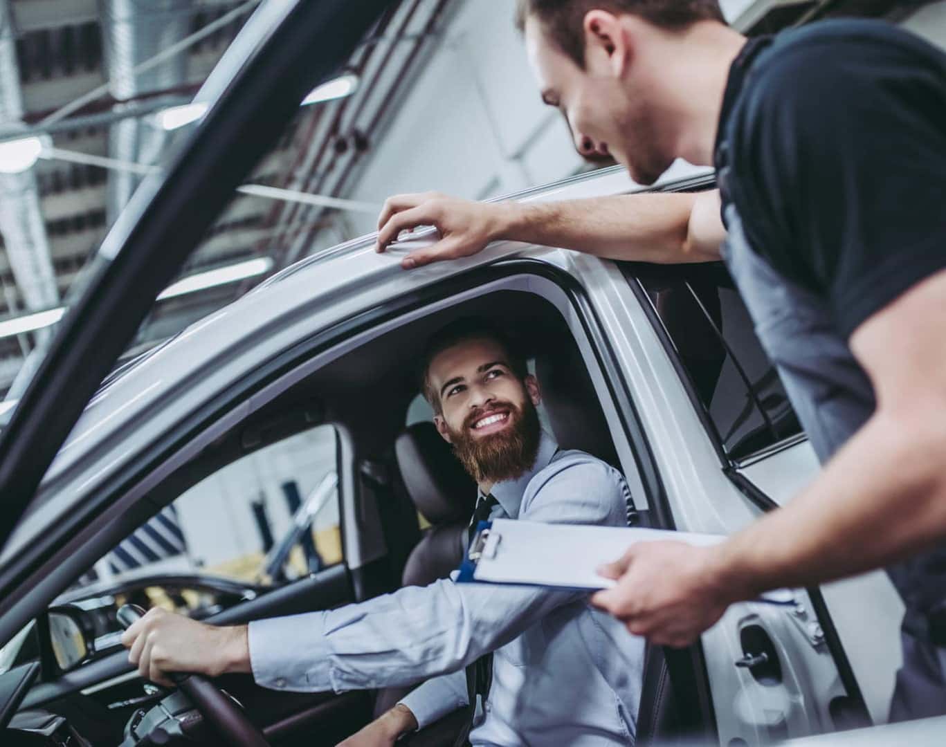 A service advisor speaking with a customer seated in the driver’s seat of a vehicle inside a dealership service center.