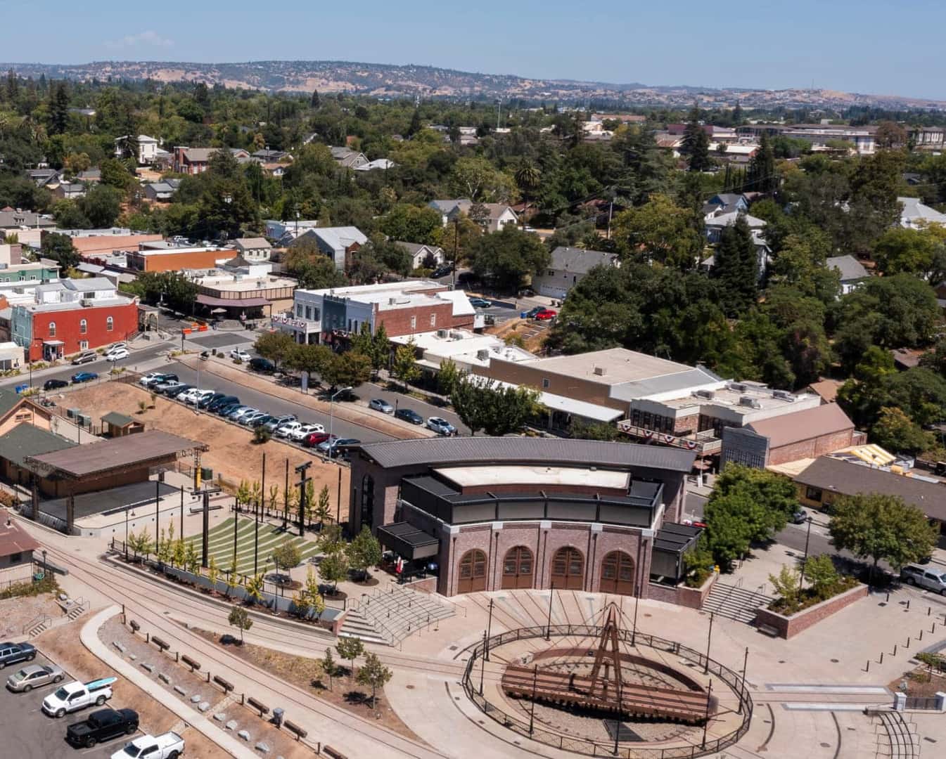 An aerial view of a downtown district featuring a central plaza, historic-style commercial buildings, parking areas, and surrounding residential neighborhoods.