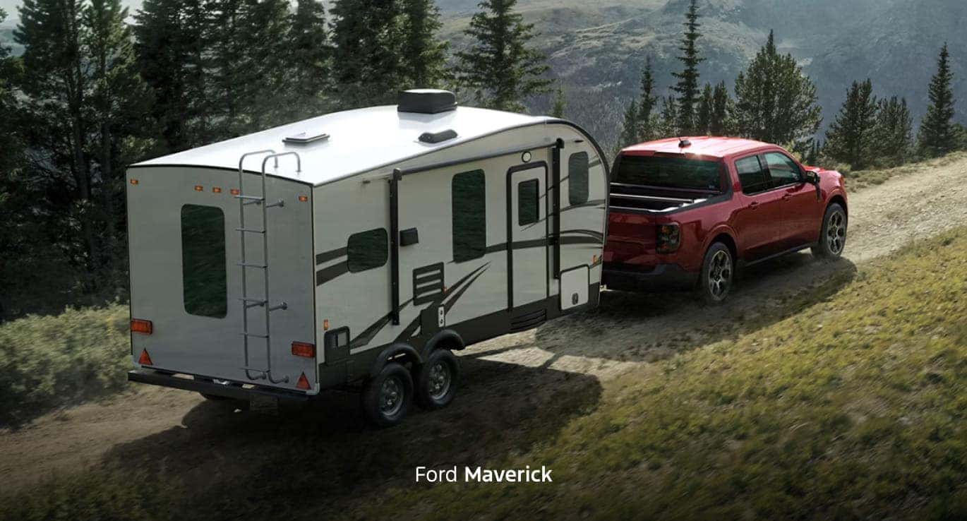 Red Ford Maverick towing a large travel trailer along a rugged mountain trail surrounded by pine trees.