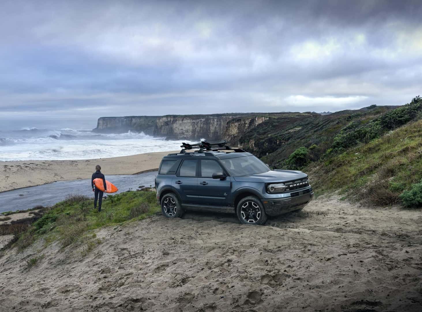 Ford Bronco parked on the beach with surfer looking out at the water