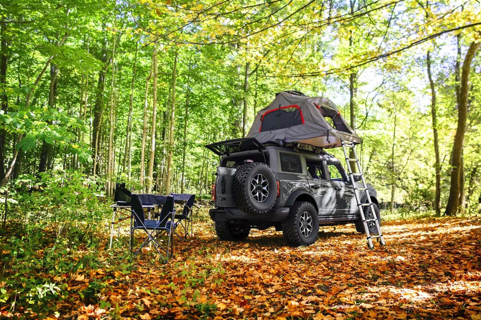 Ford Bronco Sport Parked in the woods with a tent on top of vehicle with later attached and a foldout table and chairs next to it