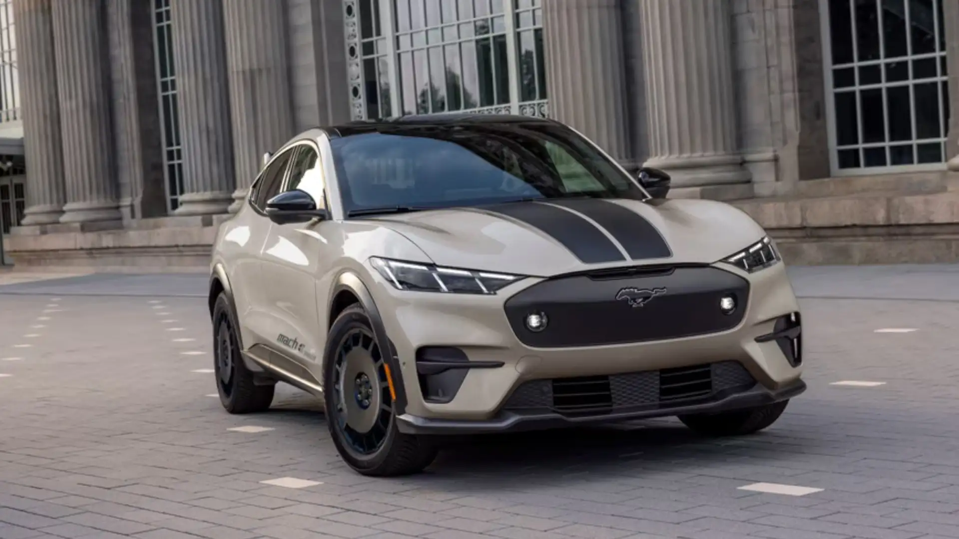 2025 Ford Mustang Mach-E in beige with black hood stripe parked by tall columns, front-angle view.
