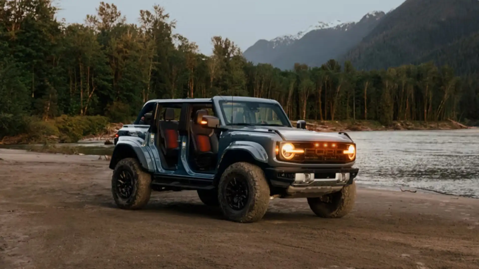 Ford Bronco with doors removed parked on a sandy riverside trail at sunset, rugged off-road stance.