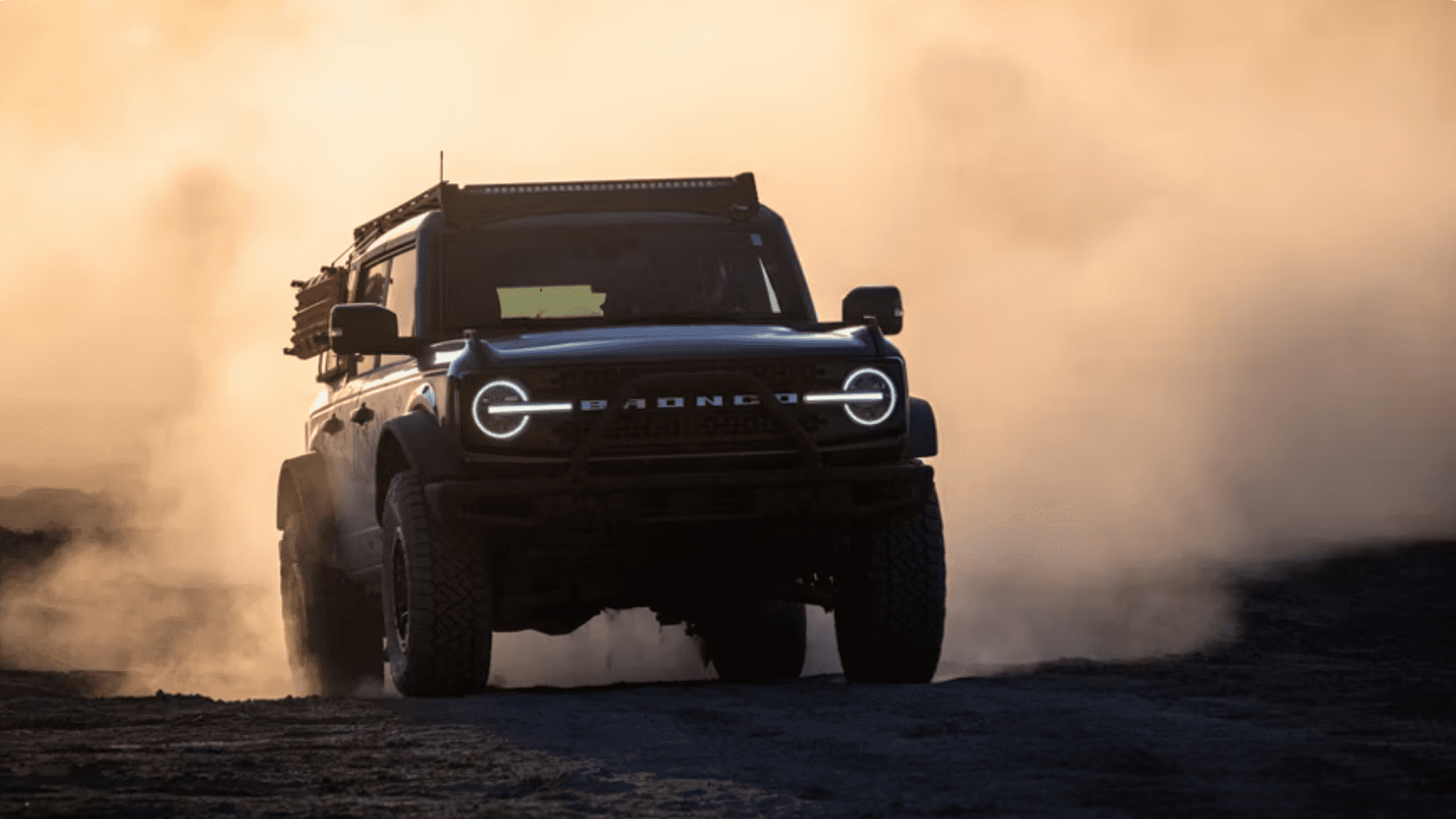 2025 Ford Bronco® off-road SUV driving through dusty terrain at sunset with LED headlights illuminated.