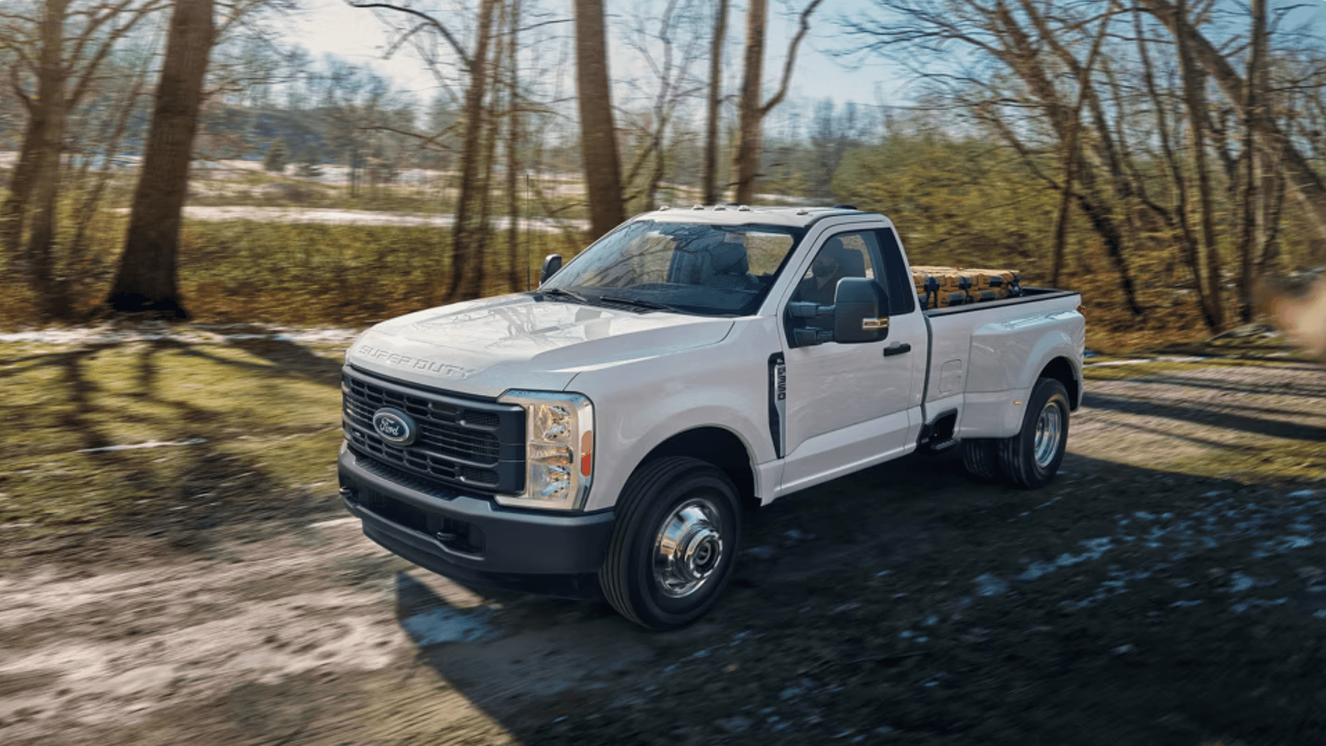 2026 Ford Super Duty® work truck driving on wooded road carrying lumber in open bed.