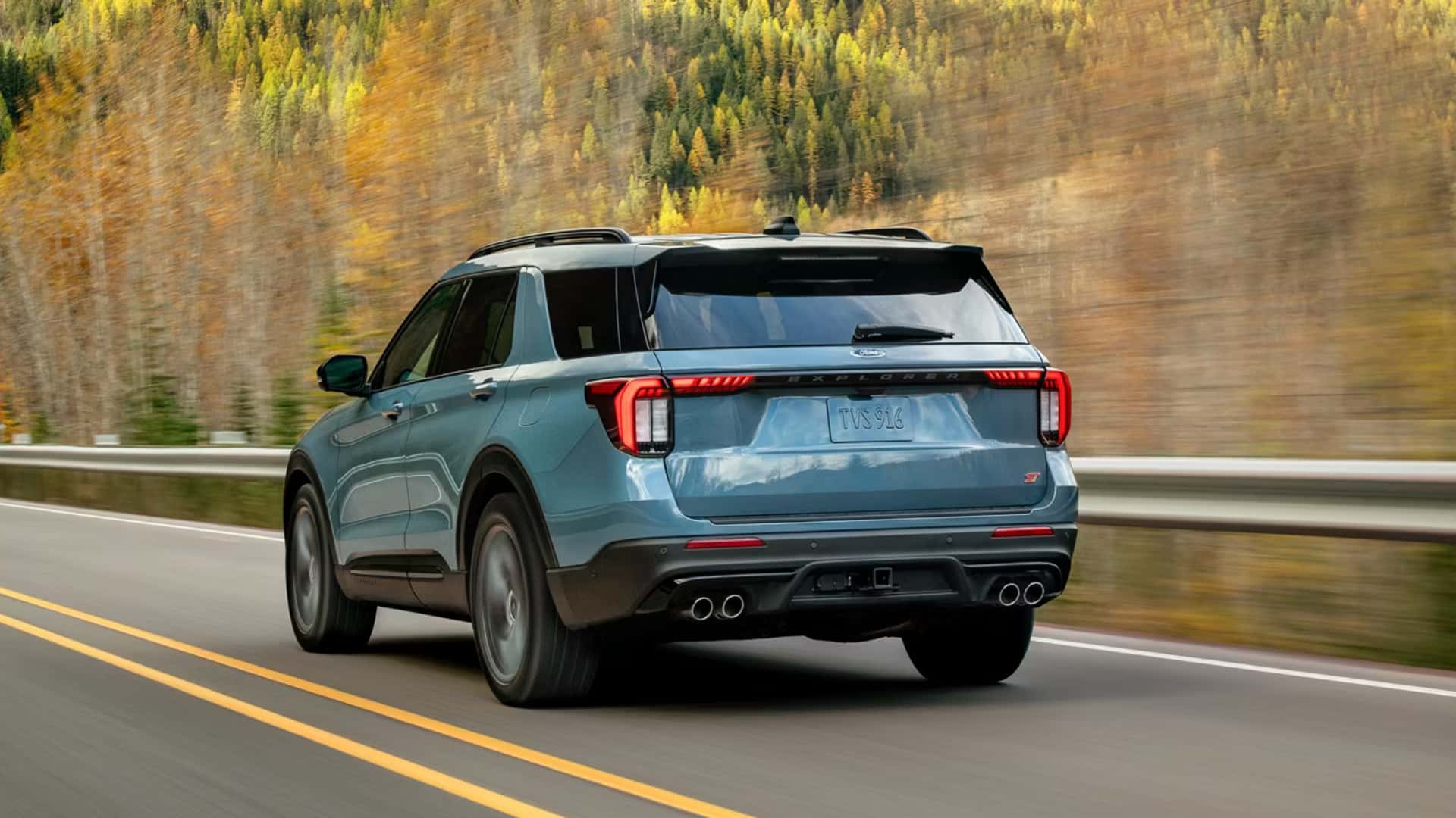 2026 Ford Explorer® SUV driving on a highway, rear three-quarter view showing taillights, roof rails, and dual exhaust.