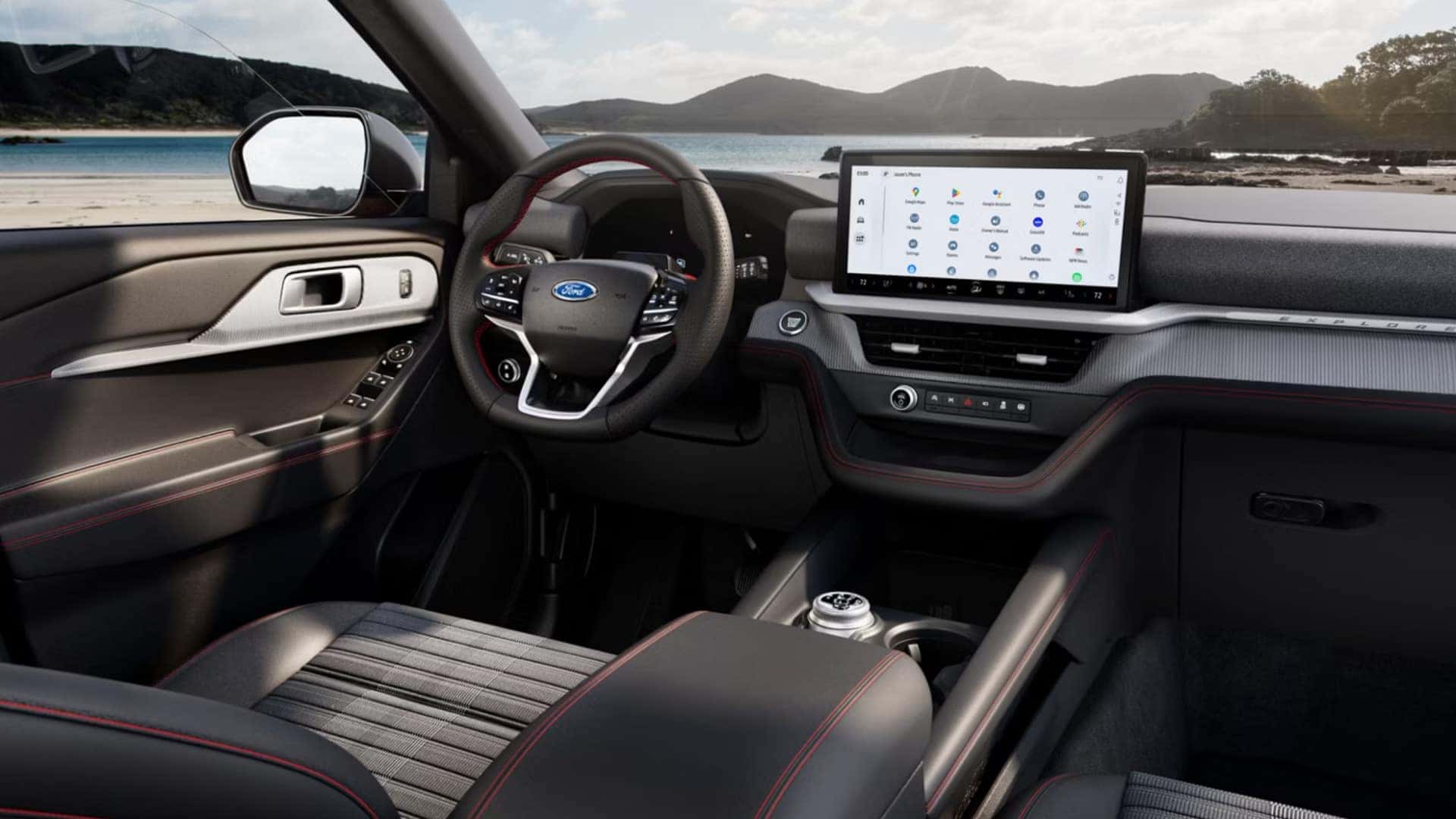 2026 Ford Explorer® cockpit view with Ford steering wheel, wide touchscreen infotainment display, and dashboard trim, with a coastal landscape visible outside.