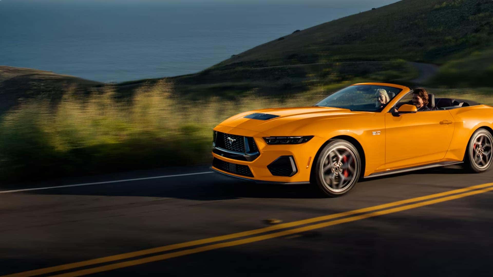 2026 Ford Mustang® convertible in orange driving on a scenic coastal road, side view with two passengers and motion blur in the background.