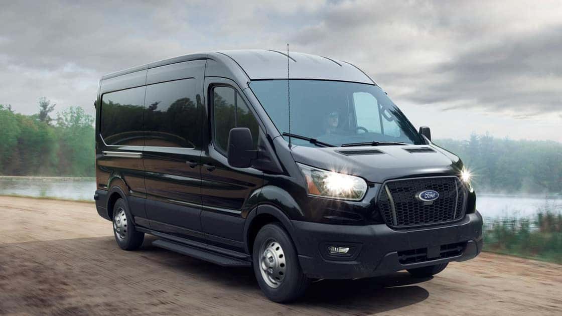 2026 Ford Transit® cargo van driving on a road with headlights on under a cloudy sky.