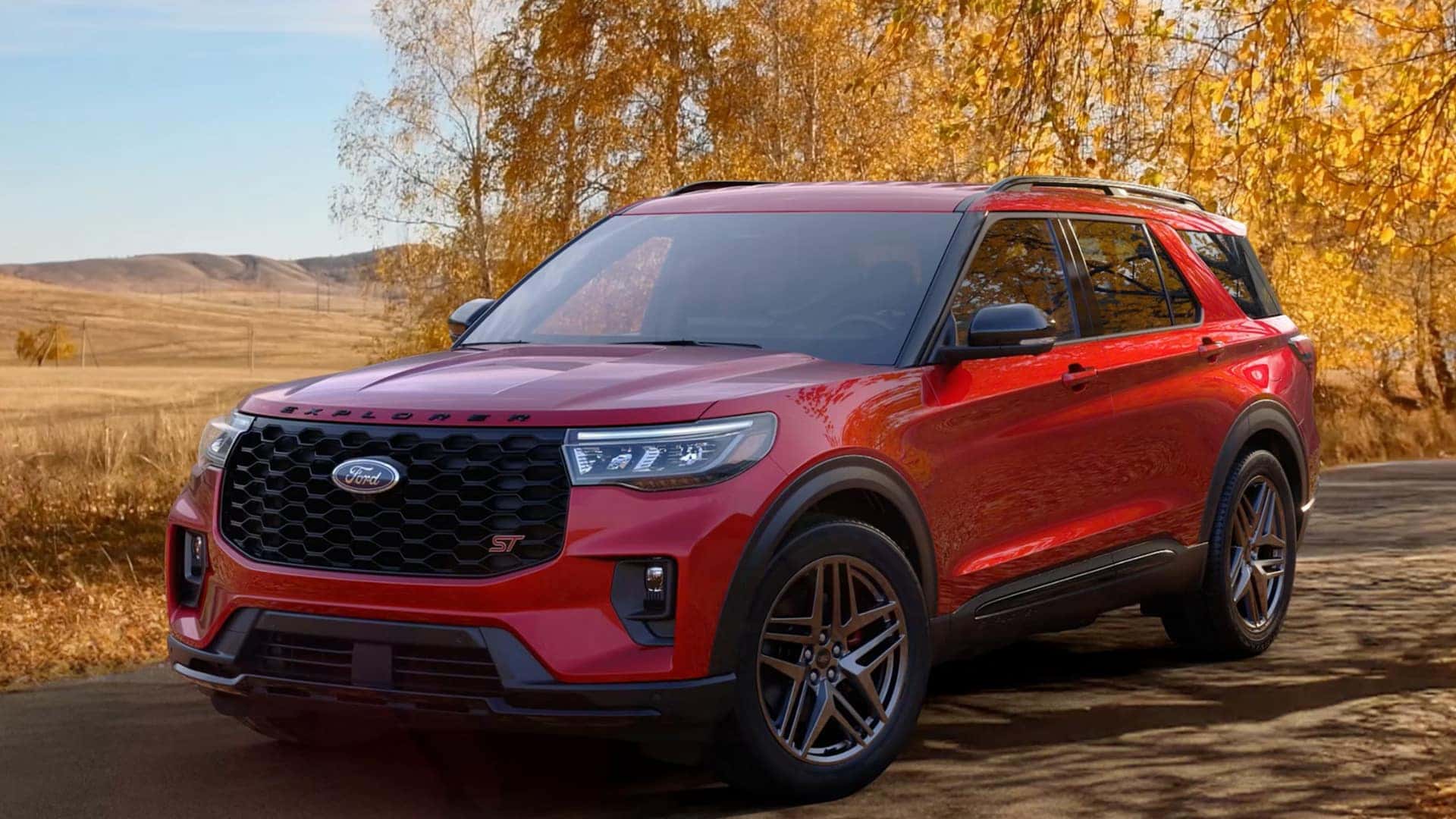 2026 Ford Explorer® red exterior parked on scenic fall road, side view showing front grille, headlights, and wheels.