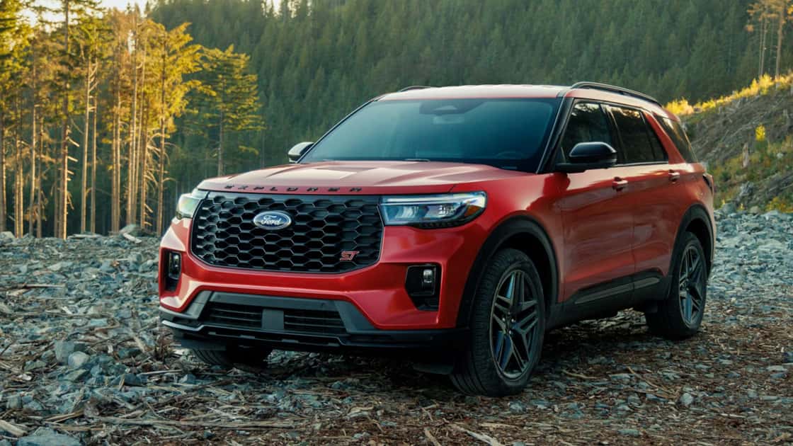 Red 2026 Ford Explorer® SUV parked on rocky terrain in a forested area, showcasing front grille and headlights.