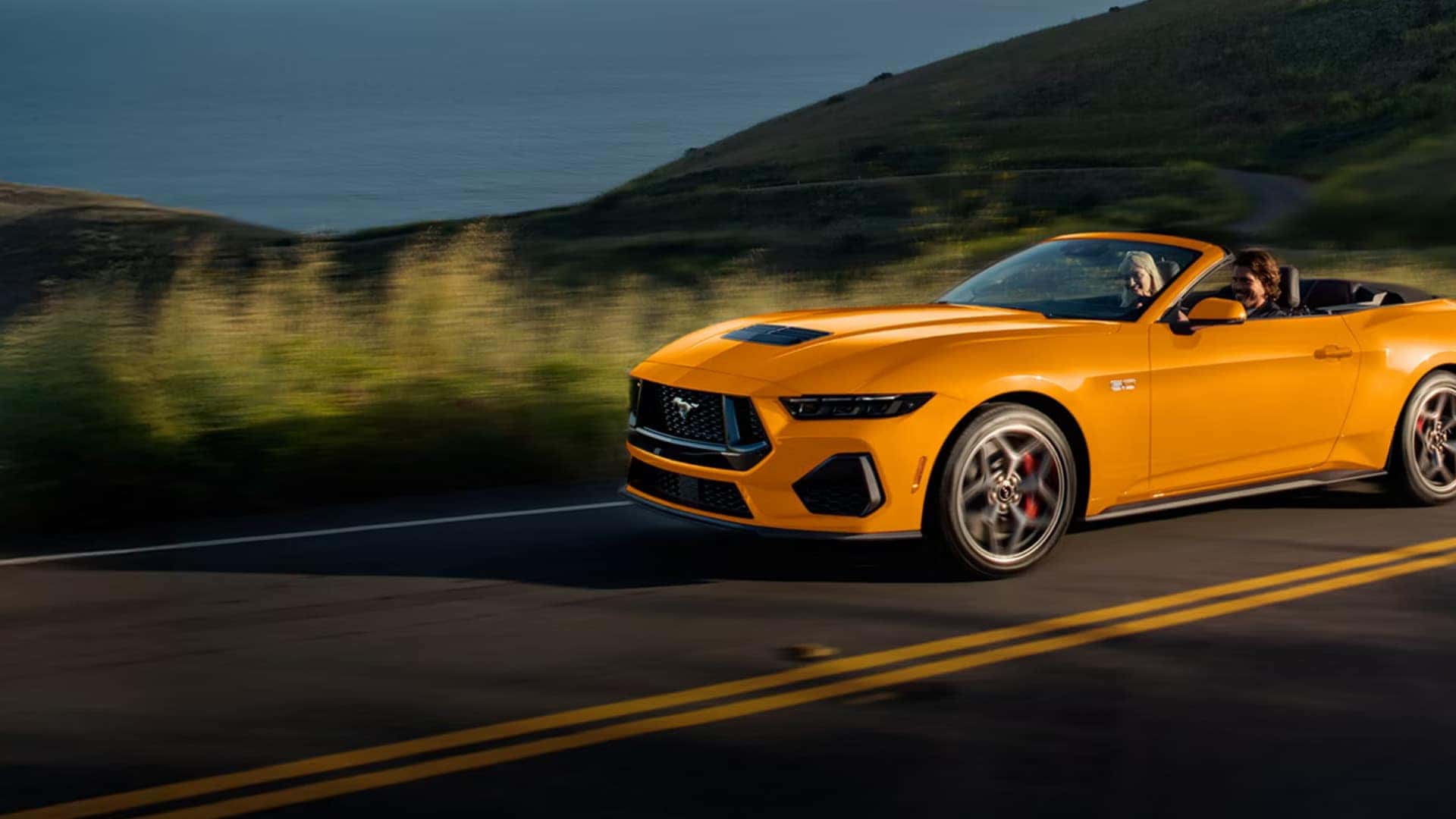 2026 Ford Mustang® yellow convertible driving on a coastal road with driver and passenger visible.