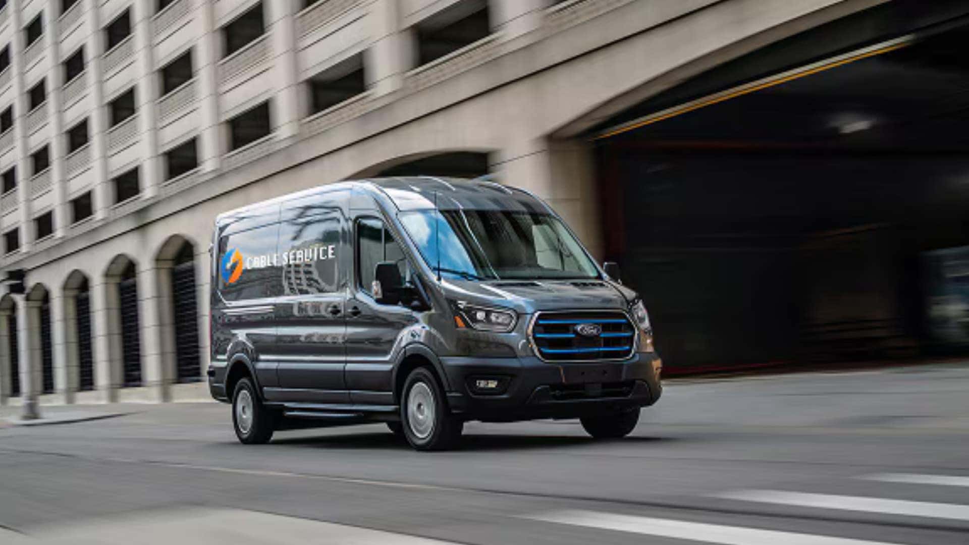 2026 Ford E-Transit™ service van driving through a city street with branded cargo body and electric front grille.