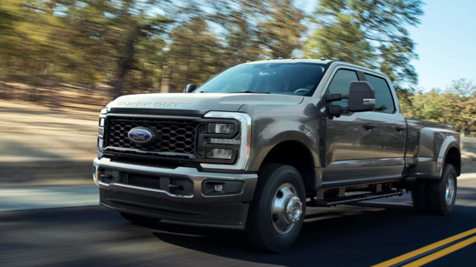 2026 Ford Super Duty® gray dually truck driving on a highway with bold front grille, towing mirrors, and heavy-duty stance.