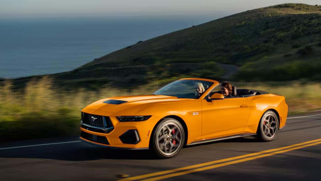 2026 Ford Mustang® yellow convertible driving on a scenic coastal road with the top down.