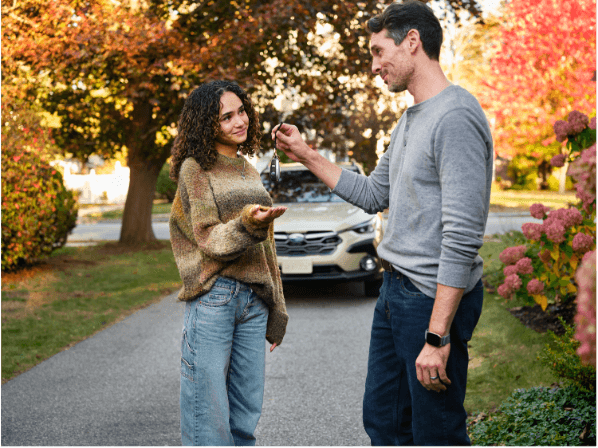 A man handing car keys to a young woman in a driveway surrounded by fall foliage, representing the trade-in process at Goode Motor Ford in Burley, ID
