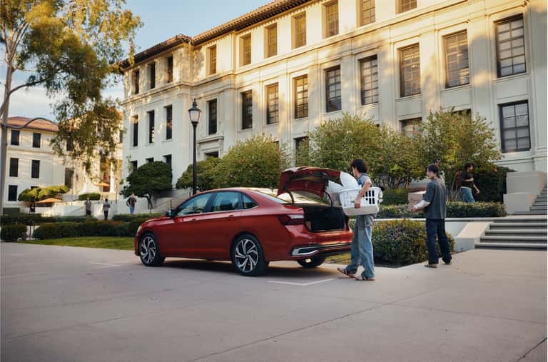 Two young adults unloading boxes and a laundry basket from the trunk of a red sedan outside a university building, showing how any vehicle can be traded in at Goode Motor Ford