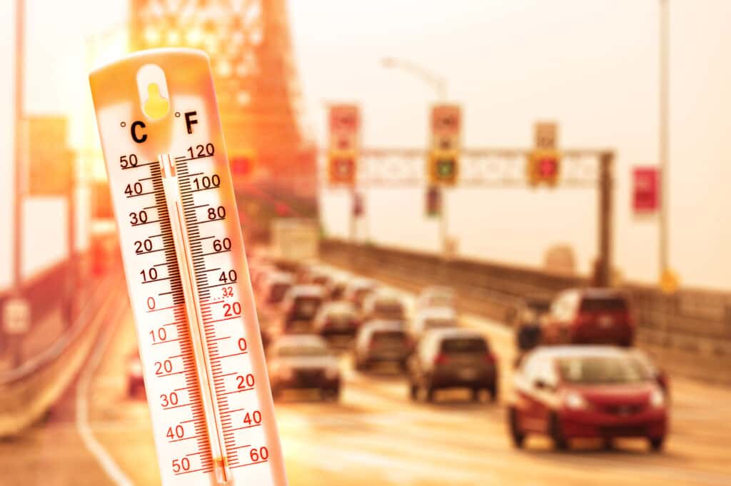 A thermometer shows a scorching hot day with cars in traffic behind it.