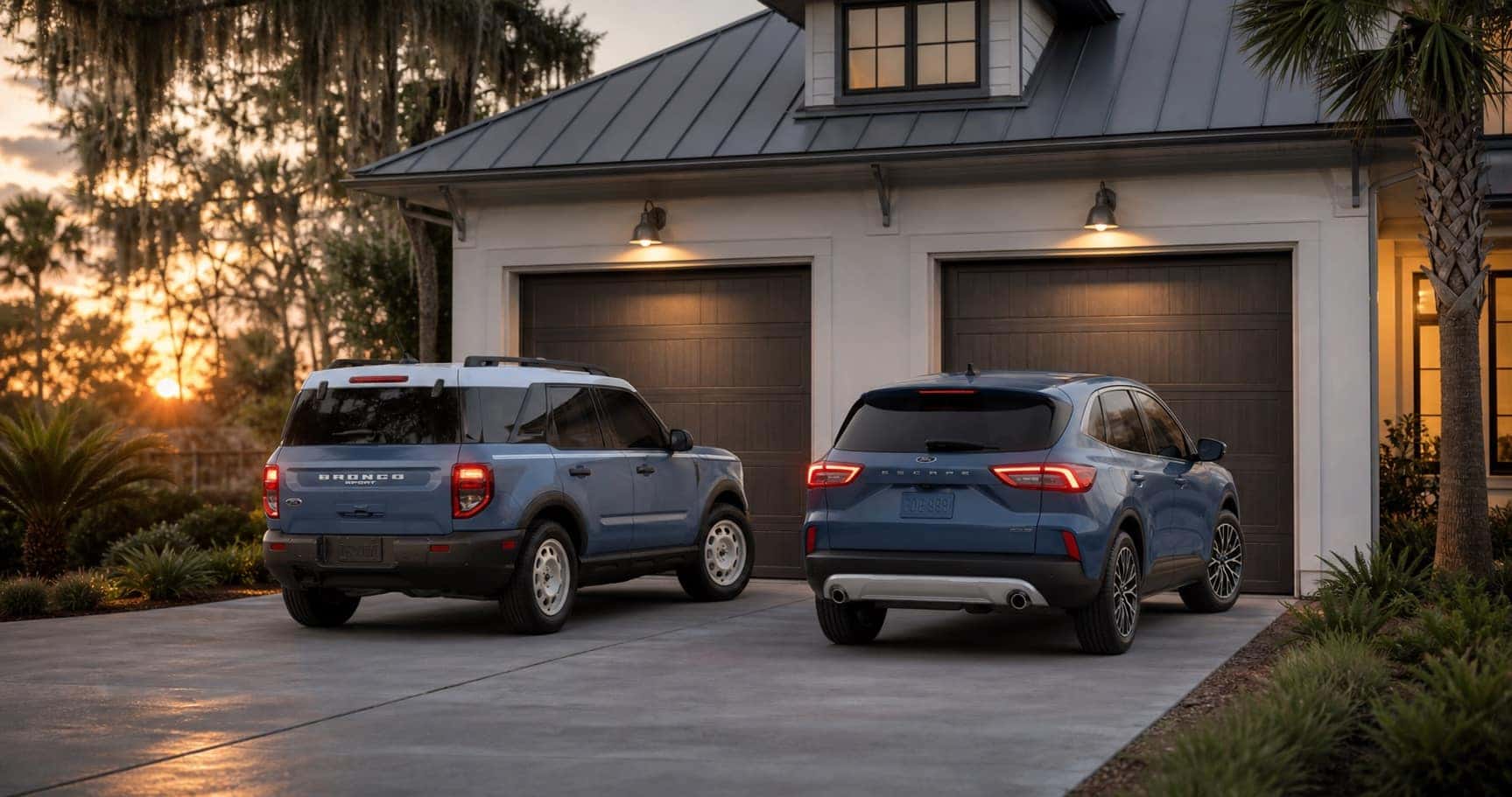 Blue Ford Bronco Sport and blue Ford Escape parked side by side in driveway at sunset in front of coastal home highlighting rear styling and design differences.