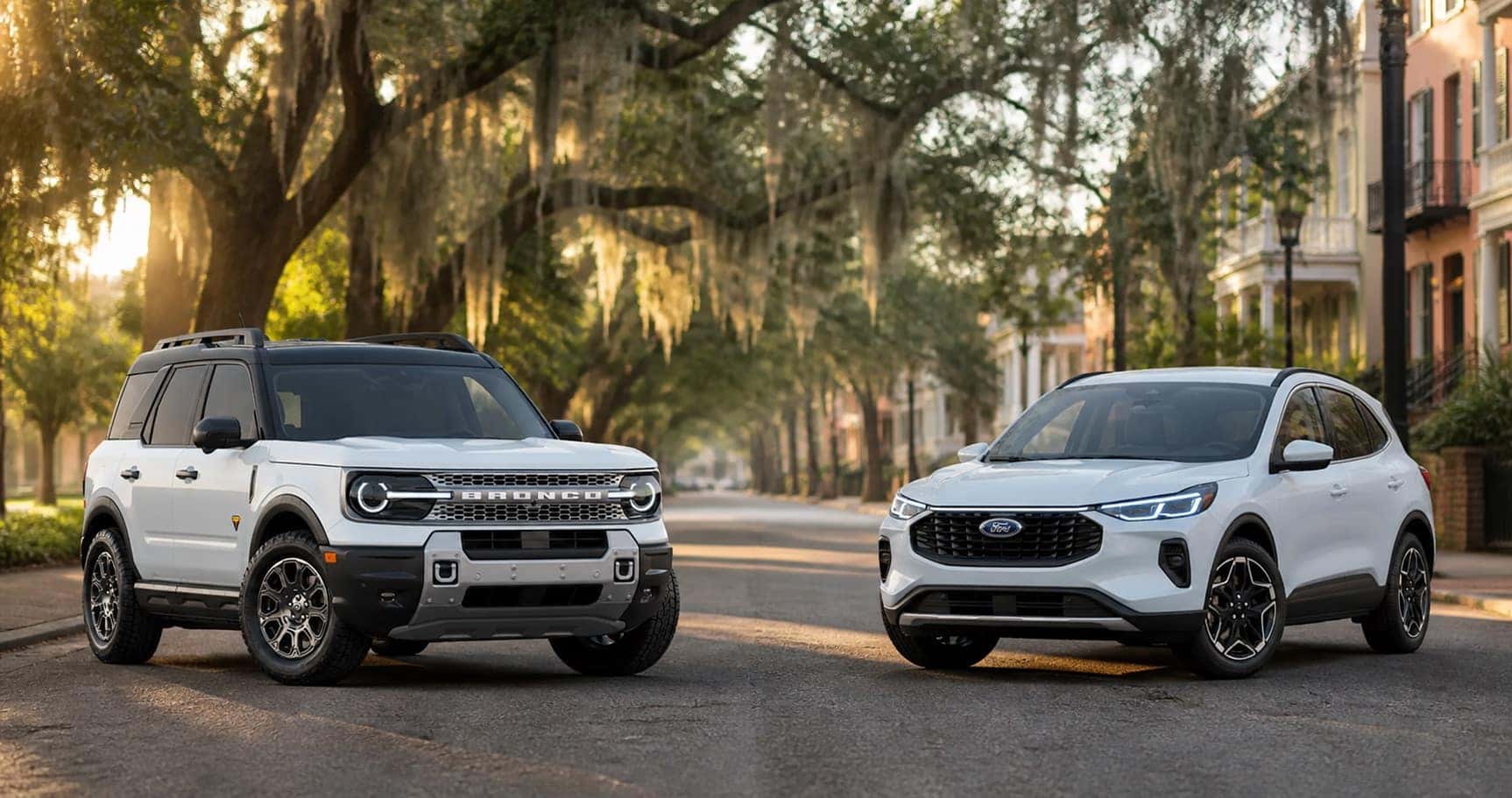 White Ford Bronco Sport and white Ford Escape parked on tree lined street under Spanish moss at sunrise showcasing front grille designs and trim level styling.