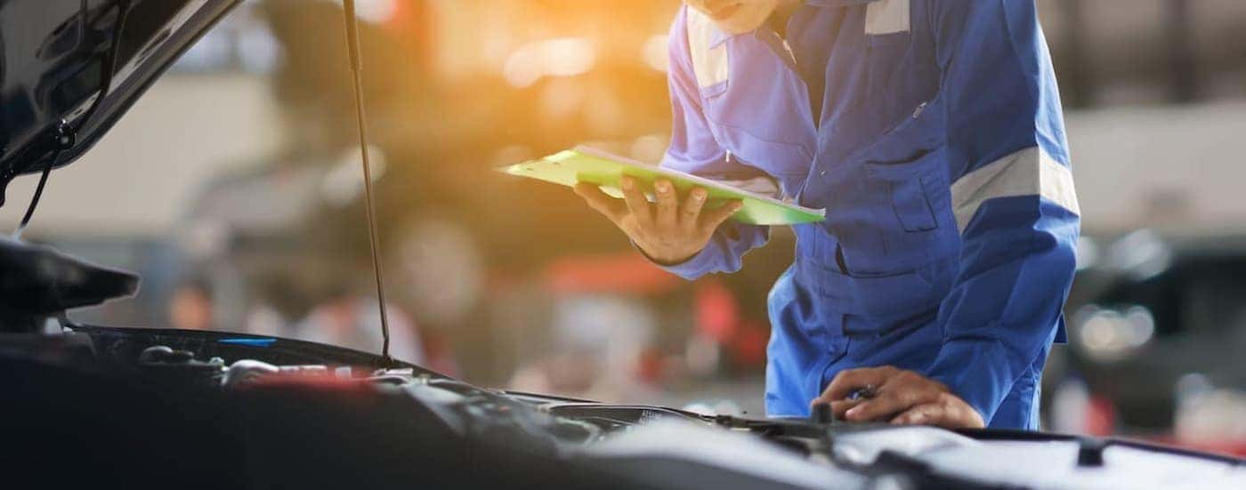 A mechanic is shown holding a clipboard near a vehicles engine bay.