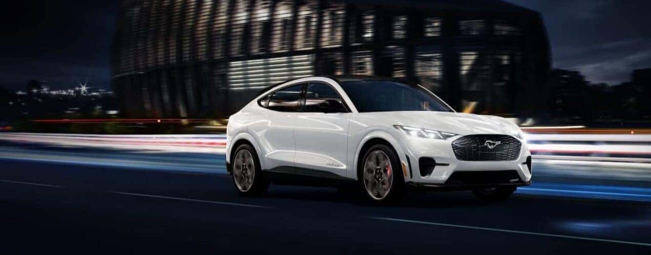 A white 2025 Ford Mustang Mach-E is shown driving near a stadium.