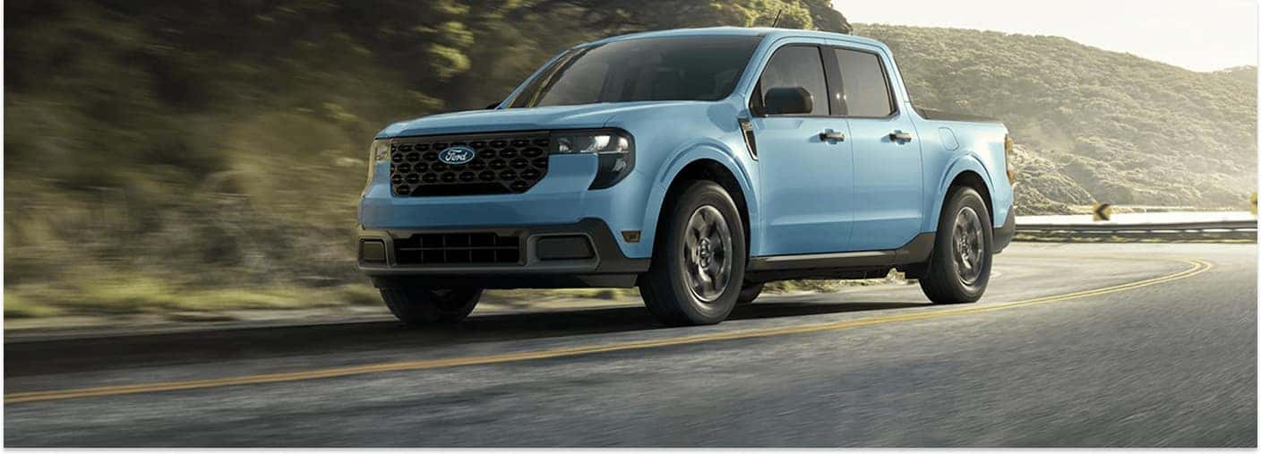 > Front three-quarter view of a light blue 2026 Ford Maverick pickup truck driving through a curve on an asphalt road with lush green hills in the background.