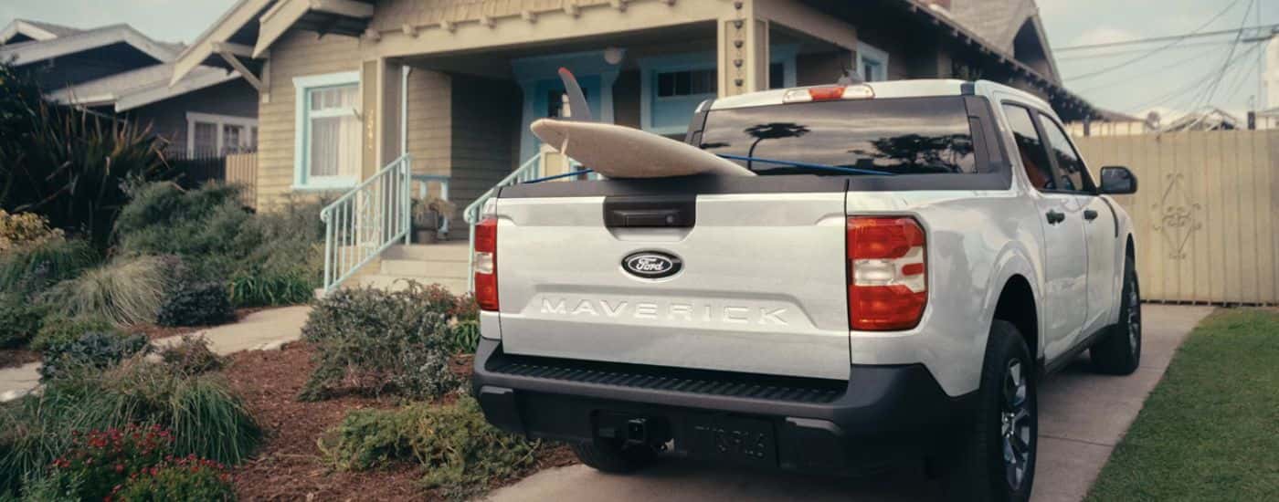A white 2026 Ford Maverick parked in a driveway with a surfboard in truck bed.