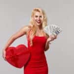 A young woman in a red dress holds in her hands cash banknotes money, dollars and a heart shaped gift box. Valentines Day and March 8