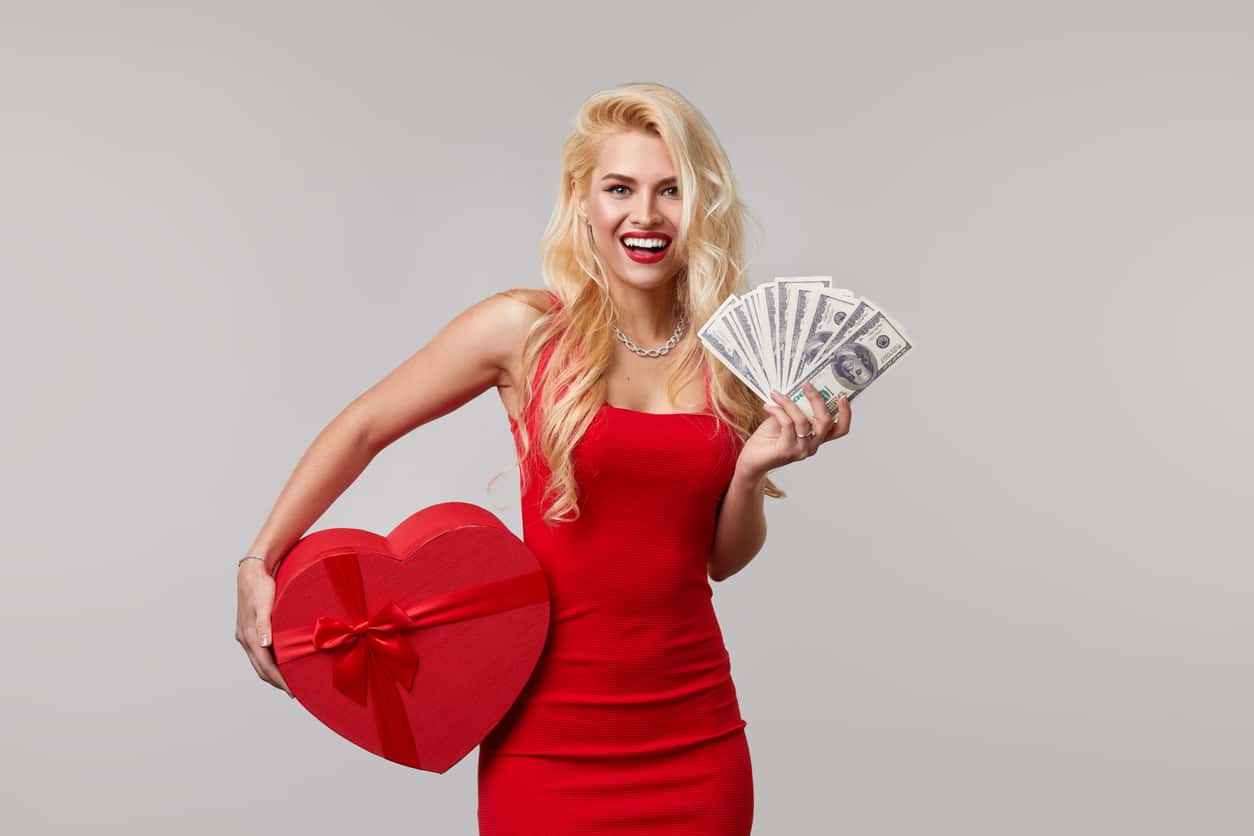 A young woman in a red dress holds in her hands cash banknotes money, dollars and a heart shaped gift box. Valentines Day and March 8