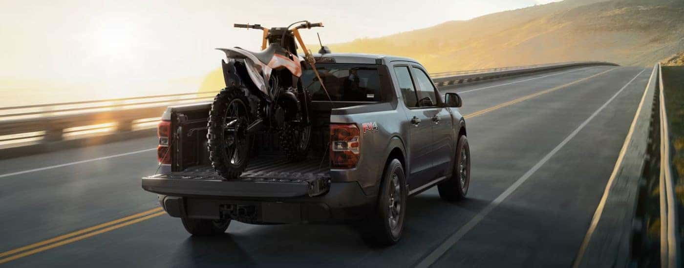 Grey 2026 Ford Maverick driving down the road with a dirt bike in the bed