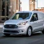 A white Ford Transit Connect is shown driving on a road.