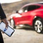 A close up shows a person holding a phone with the FordPass Connect app in front of a red 2022 Ford Mustang-Mach-E.