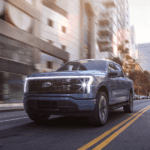 A light blue 2022 Ford F-150 Lightning is shown driving down a city street after leaving a Zionsville Ford dealer.