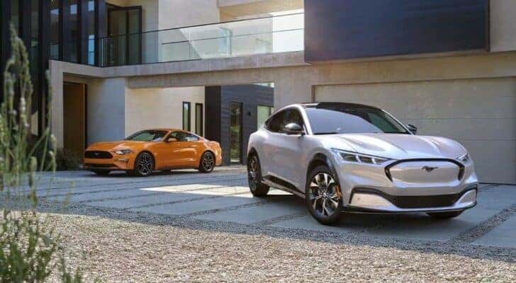 An orange 2022 Ford Mustang and white Mustang Mach-E are shown parked on the driveway of a modern house.