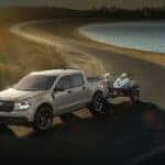 A silver 2024 Ford Maverick is shown towing a trailer to a car dealership.