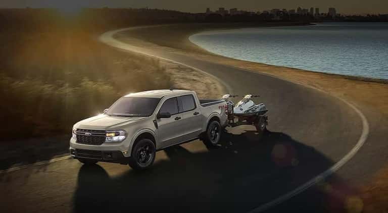 A silver 2024 Ford Maverick is shown towing a trailer to a car dealership.