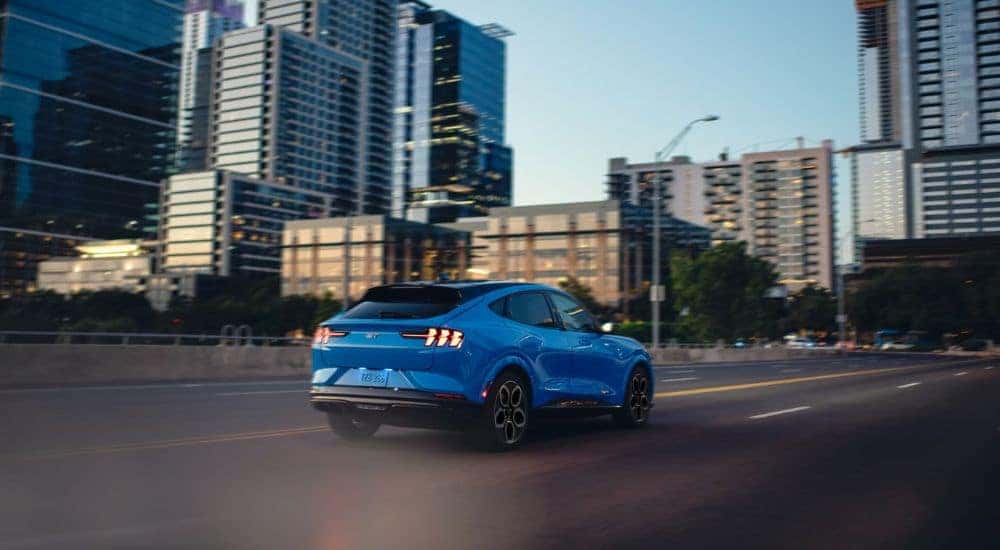 A blue 2024 Ford Mustang Mach-E for sale near Carmel is shown driving past buildings.