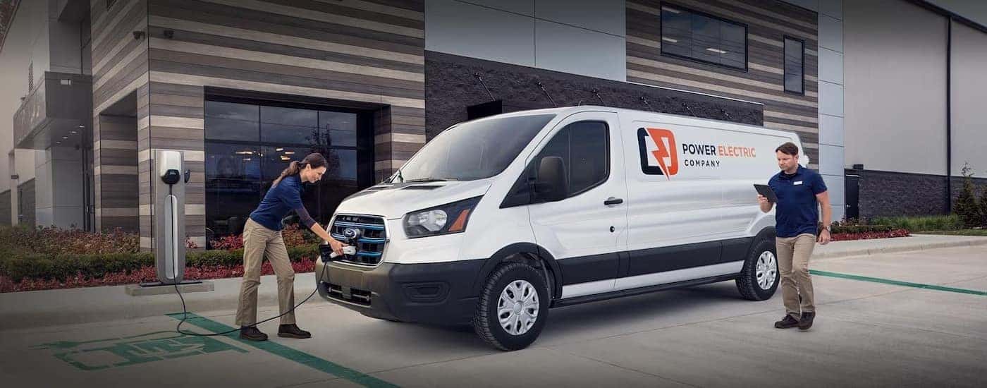 A white 2023 Ford E-Transit van is shown charging near two people.