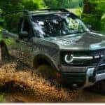 Front three-quarter view of a gray 2026 Ford Bronco Sport driving through a muddy puddle on a forest trail.