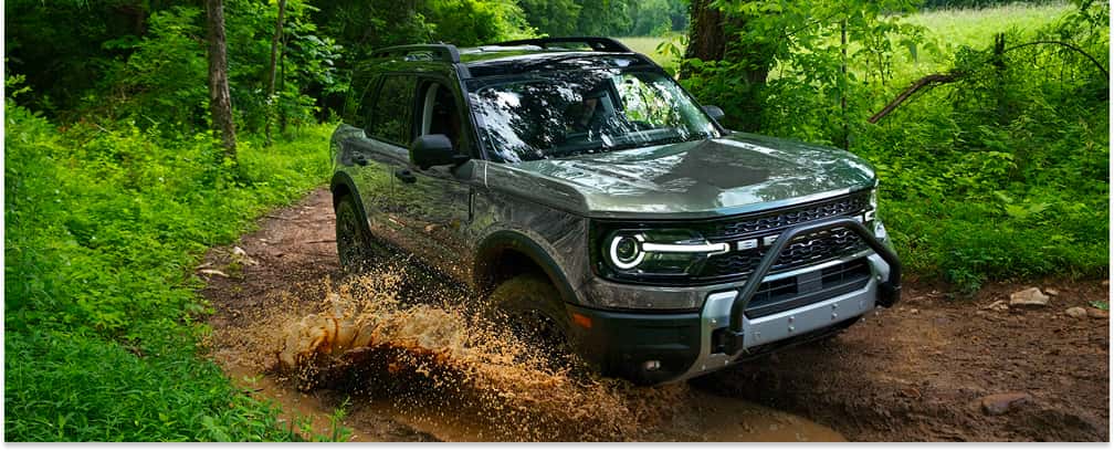 Front three-quarter view of a gray 2026 Ford Bronco Sport driving through a muddy puddle on a forest trail.
