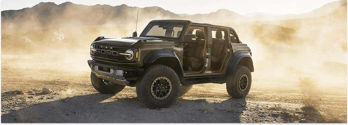 Front three-quarter view of a green 2026 Ford Bronco Raptor driving with doors off on a dusty desert trail.