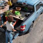 A woman is shown putting gardening supplies into the bed of a blue 2023 Ford Maverick.